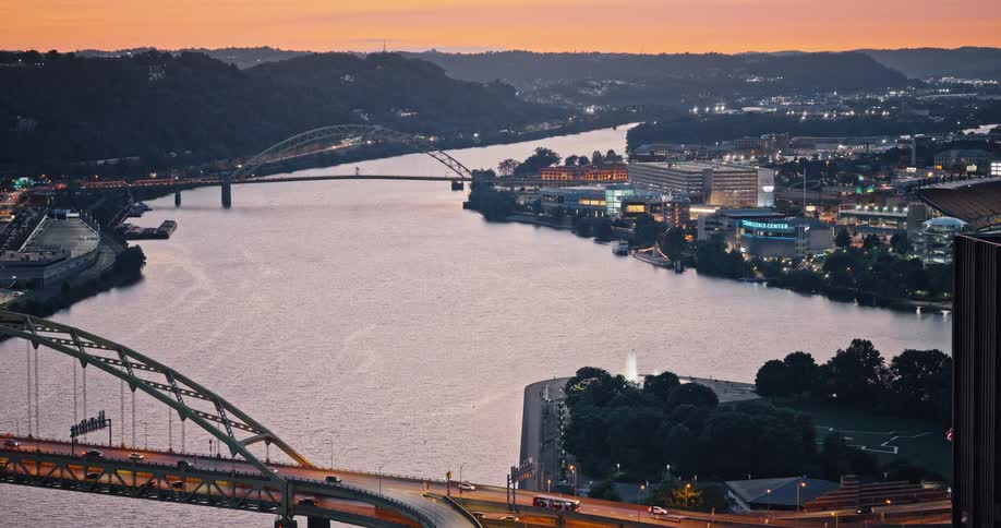 Aerial view of the city of Pittsburgh at dusk, showing bridges crossing the river as traffic flows and lights twinkle