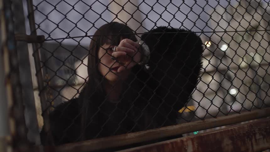 A pensive young woman in a black coat gazes through a chain-link fence at night in a moody urban setting