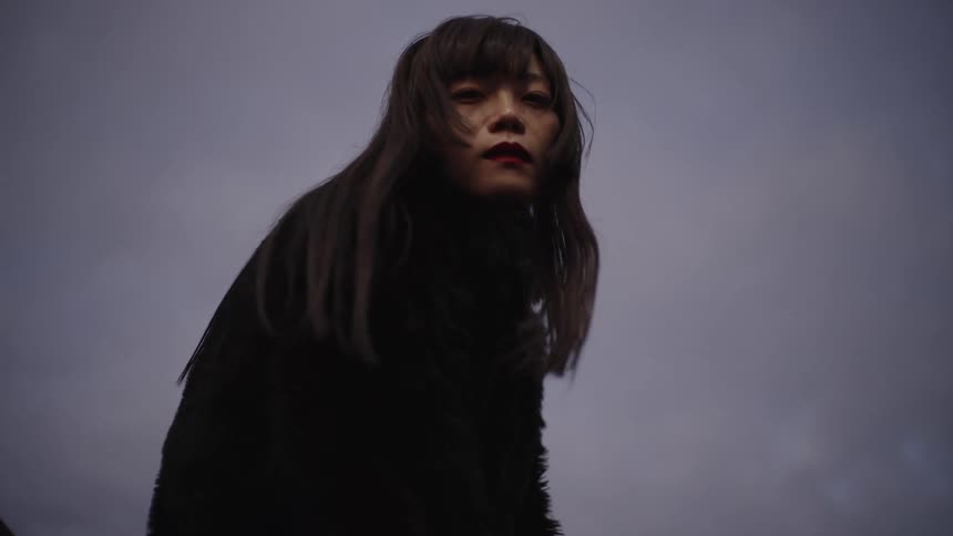 A mysterious woman with an intense gaze looks down at the camera against a dark, moody sky