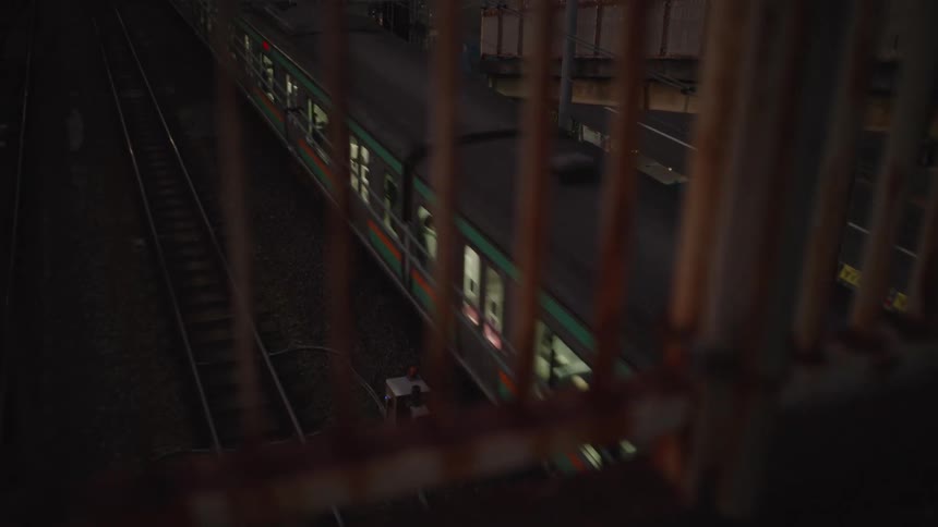 A high-angle rack focus shot reveals a commuter train passing on the tracks below, viewed through a rusty fence at night