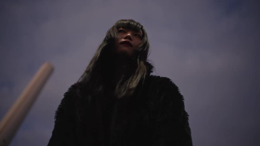 From a low angle, a young Asian woman in a black coat raises her gaze to the twilight sky