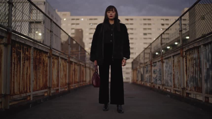 A stylish Japanese woman in an all-black outfit poses on a rusty urban bridge at dusk