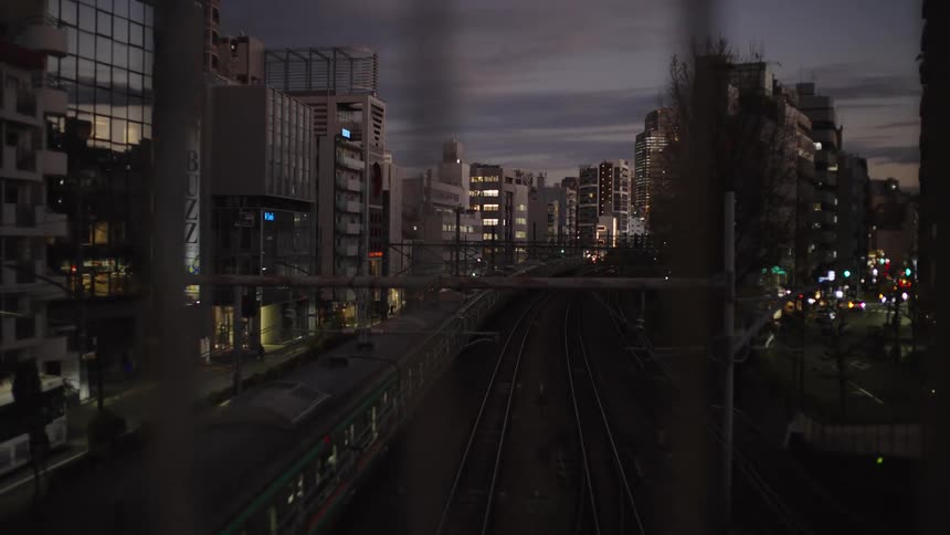 A train departs through a bustling city in Tokyo, Japan at dusk, as viewed from behind a fence