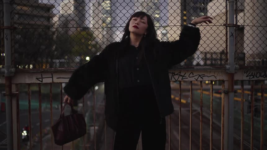 A stylish Asian woman in all black closes her eyes and embraces the city air while standing on an urban overpass at dusk