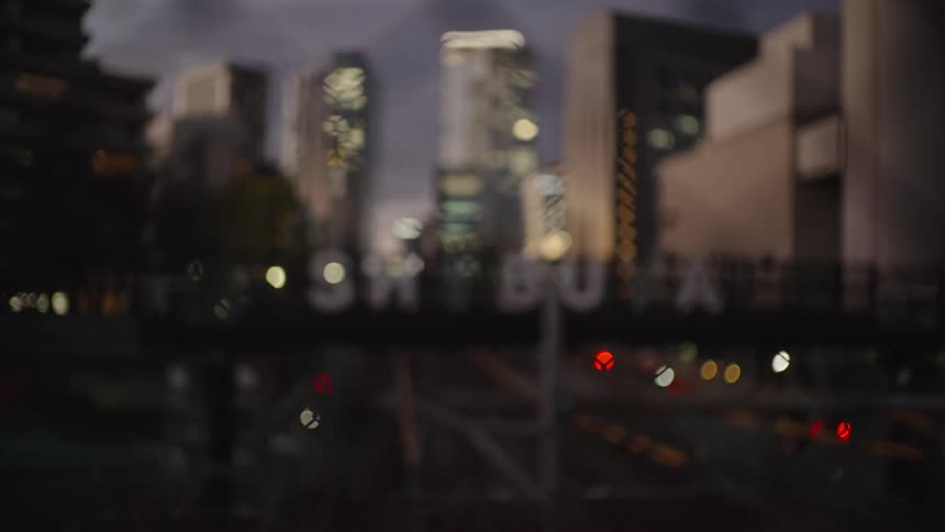 A slow rack focus from a fence reveals the Shibuya city sign over train tracks with skyscrapers in the background at dusk