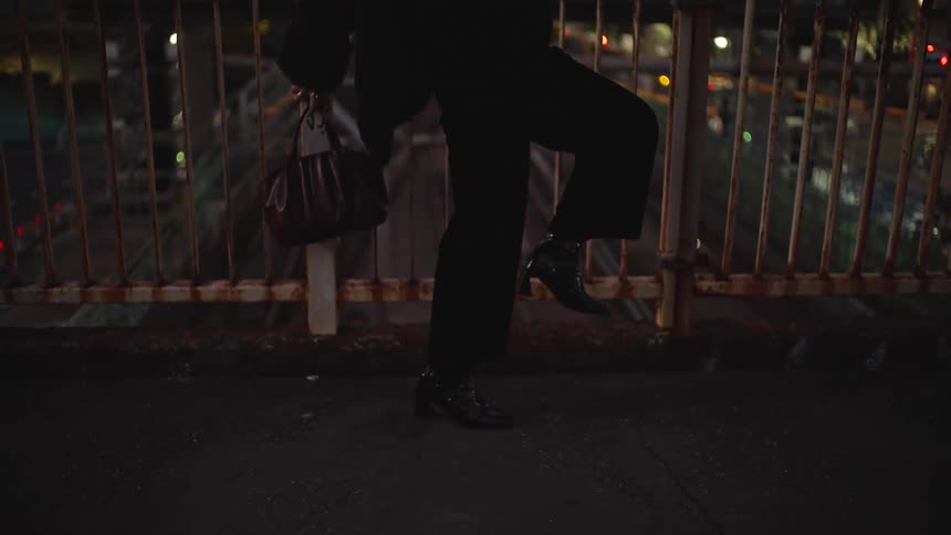 A person in black heeled boots strikes a pose on a gritty city overpass at night, with traffic lights glowing in the background