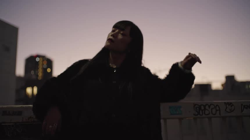 A cool and confident woman in a black fur coat poses against the city skyline at dusk in slow motion