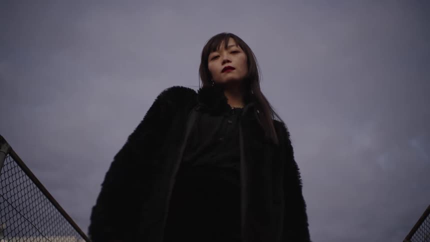 A low-angle shot captures a stylish Asian woman in a black fur coat looking down menacingly at the camera under a gloomy sky