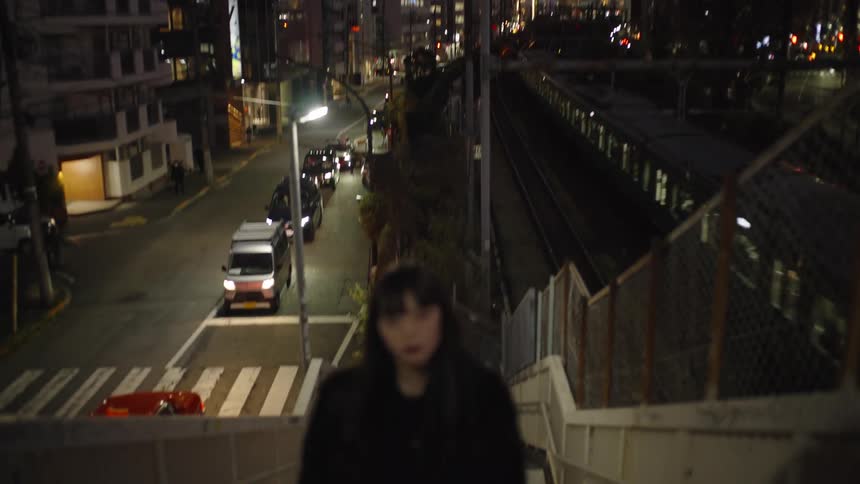 A Japanese woman walks up a set of stairs at night with a bustling city and a passing train in the background