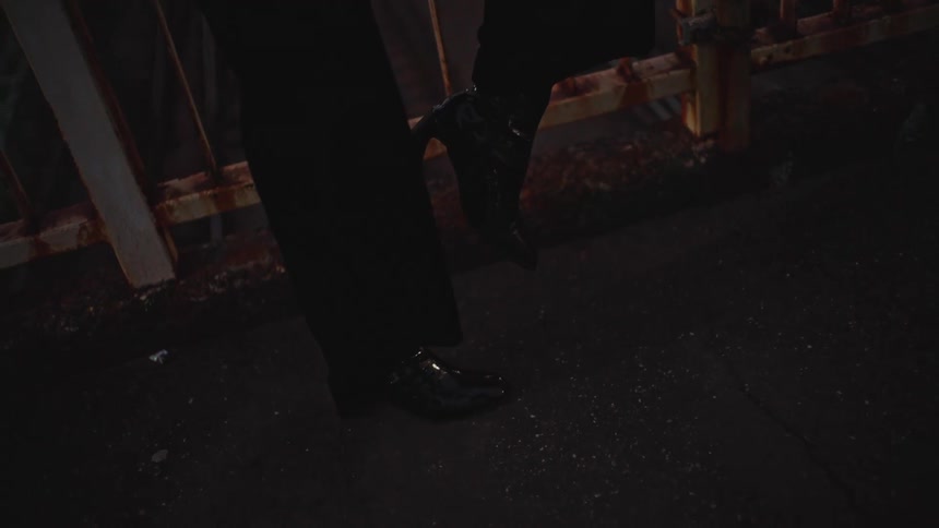 Close up on the feet of a man and woman wearing shiny black shoes, standing close together at night by a rusty fence