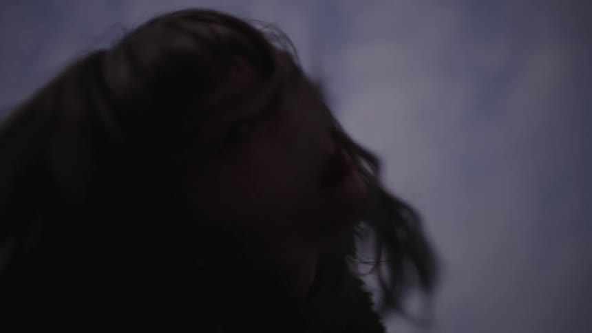 The hair of a mysterious young woman blows in the wind in slow motion, shot from a low angle against a dim, cloudy sky