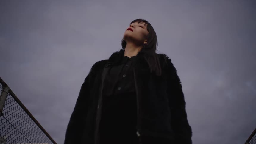 From a low angle, a fashionable woman in all black looks up at the gloomy sky, then confidently stares down at the camera