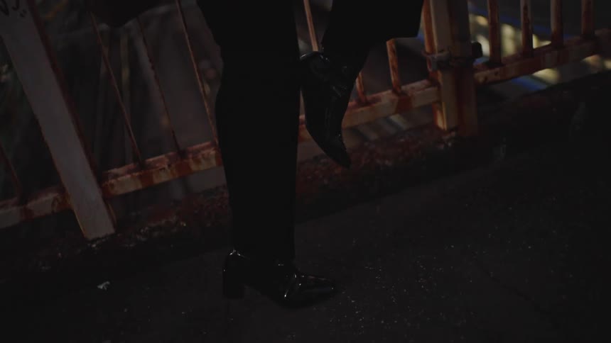 A close-up shot of a person's shiny black heeled shoes as they stand on a dark city overpass at night