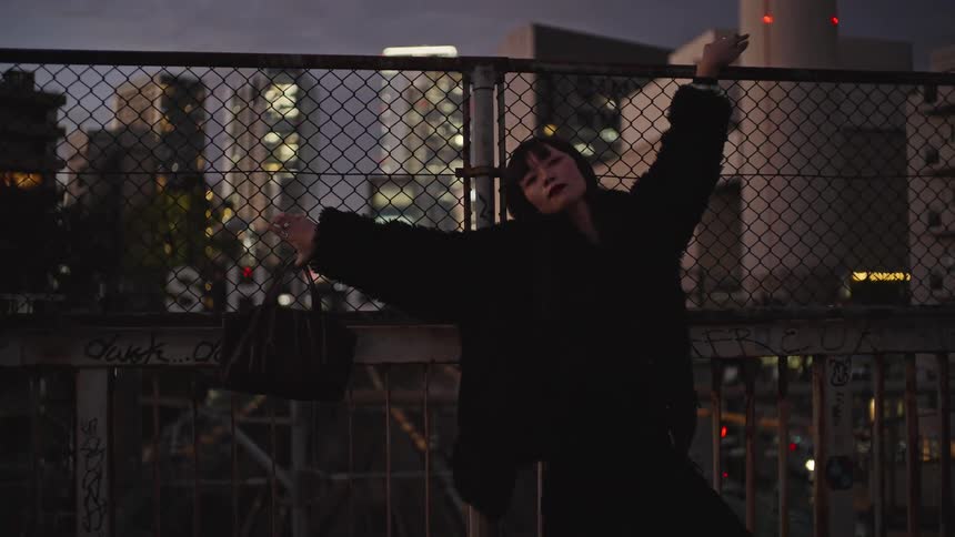 A stylish woman in a black jacket poses on an urban bridge in Shibuya, Tokyo with her arms outstretched against the cityscape at dusk