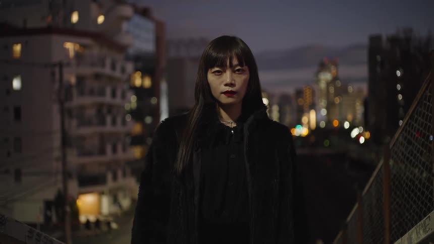 A stylish Japanese woman with long black hair and red lipstick stares intently at the camera on a city overpass at night