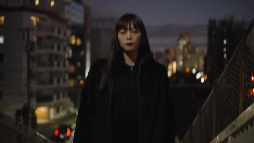 A slow tilt-up shot reveals a chic Japanese woman in a black fur coat standing on a city staircase at night