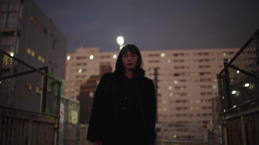 A stylish young Japanese woman walks confidently towards the camera on a city overpass at dusk