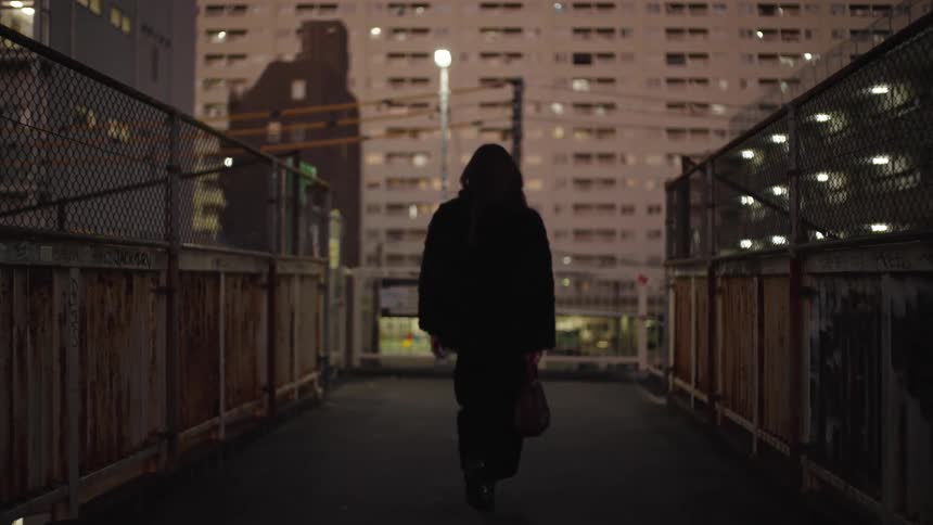 A woman in a black coat walks away from the camera down a gritty urban overpass at night towards a large apartment building