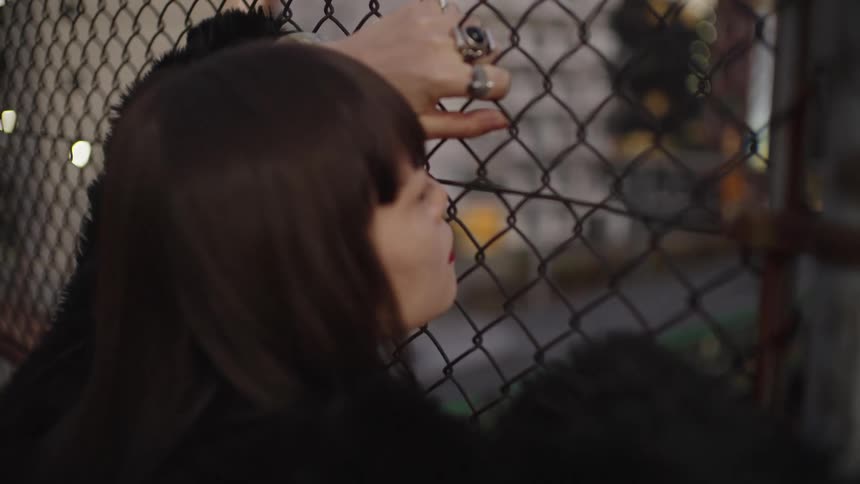 A thoughtful woman with dark hair and rings on her fingers gazes through a chain-link fence at night in an urban setting