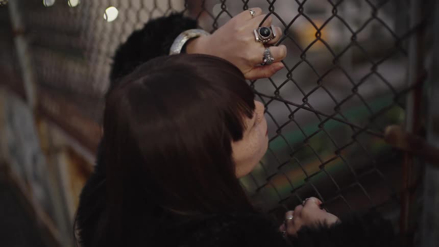 A pensive woman with dark hair and rings on her fingers looks down through a chain-link fence in a moody urban setting