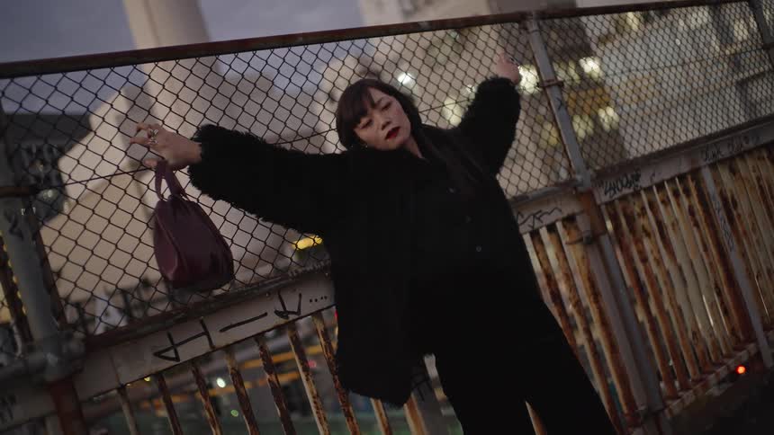 A stylish young woman in a black fur coat leans against a rusty chain-link fence on an urban overpass at dusk