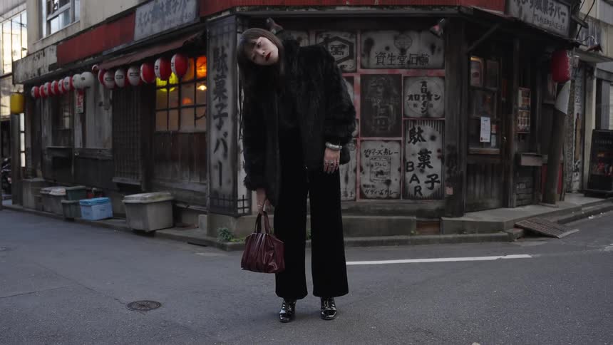 A stylish woman in a black fur coat poses with her head tilted on a corner in a quiet Japanese street