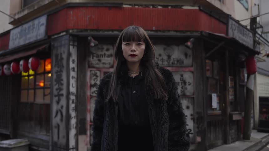 A portrait of a stylish Japanese woman in a black fur coat standing still on an urban street corner