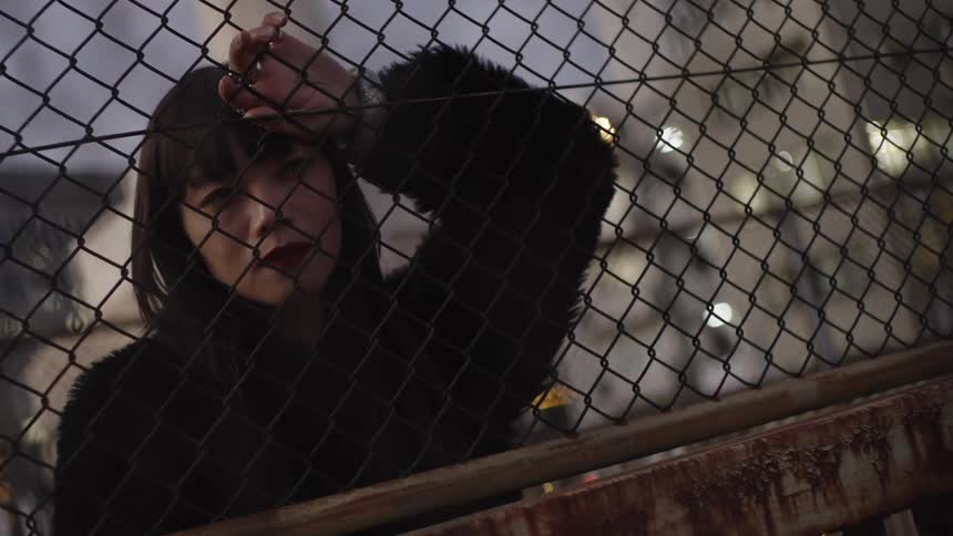 A melancholic young woman in a black coat looks through a chain-link fence at dusk with blurry city lights behind her