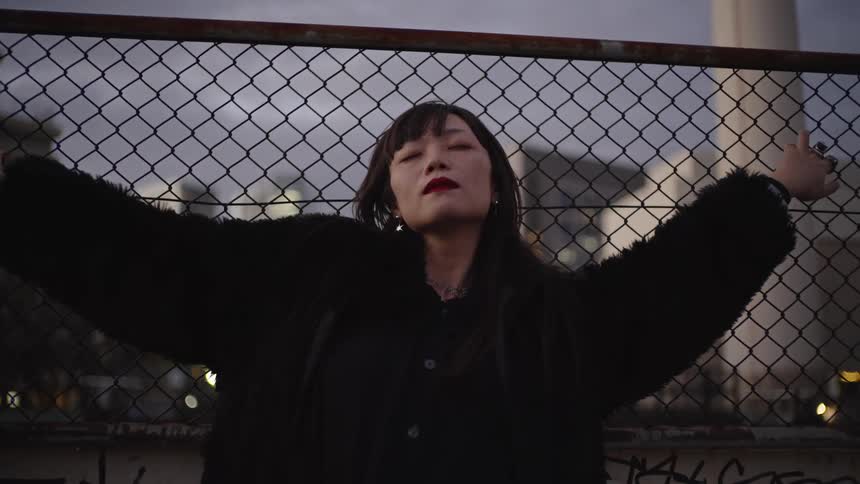 A cool Asian woman with red lipstick and a black coat leans against a chain-link fence in the city at dusk