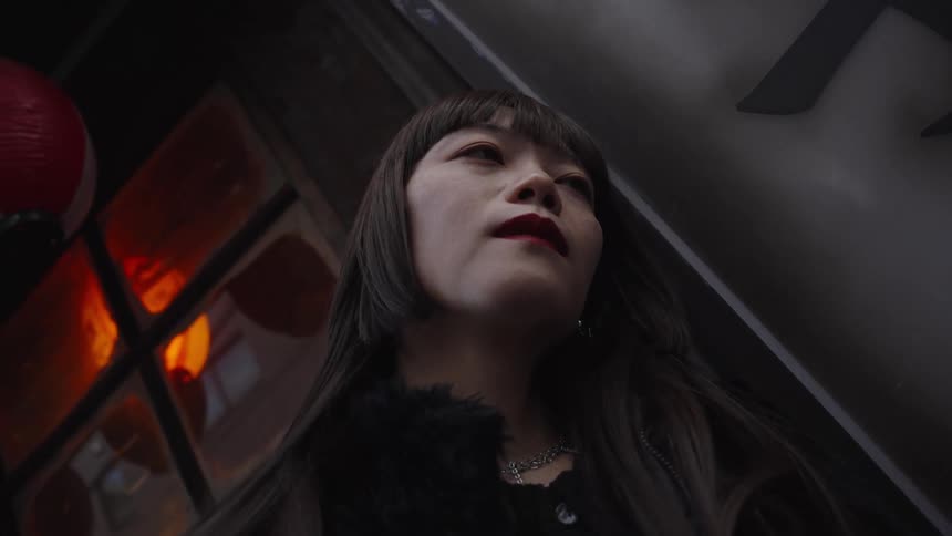 A moody low-angle shot captures a thoughtful Asian woman with long black hair standing on a city street at night