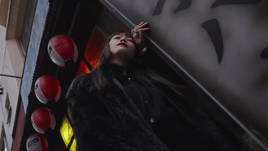 A fashionable Asian woman in a black fur coat stands pensively in a city alley next to red paper lanterns