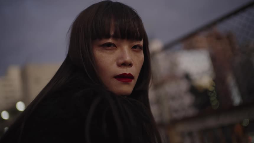 A Japanese woman with bold red lipstick gazes intensely over her shoulder in a city at dusk