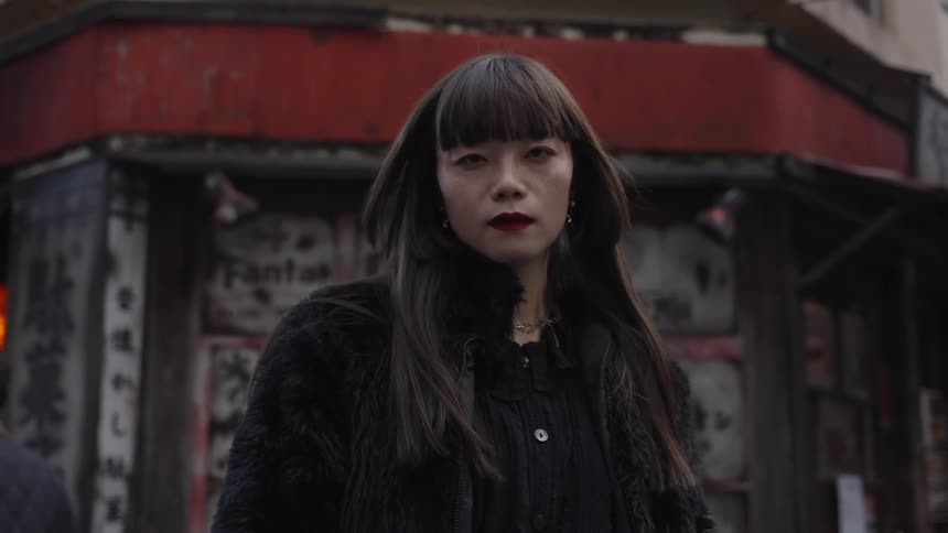 A fashionable Japanese woman with an intense gaze stands on a city street in slow motion