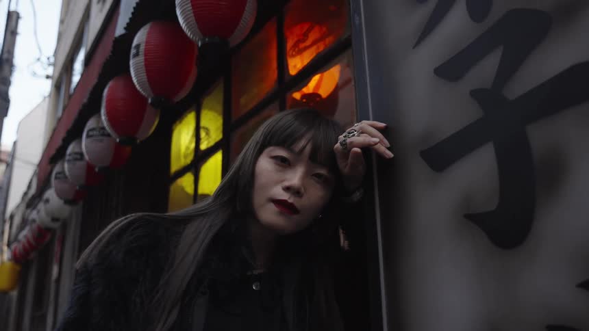 A stylish Japanese woman poses in a city alleyway next to a sign with kanji characters and traditional paper lanterns