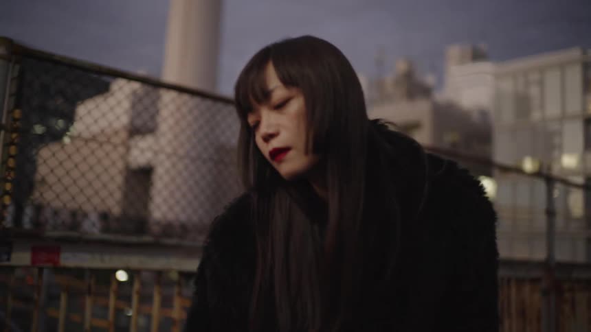 A melancholy close-up of a stylish Japanese woman in a black fur coat looking down thoughtfully in a gritty urban setting at twilight