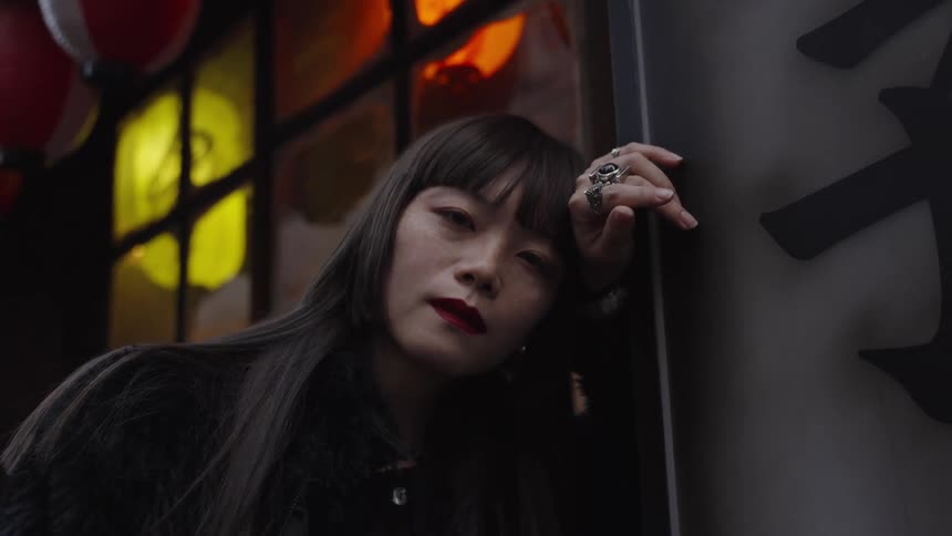 A stylish Asian woman with red lipstick gazes intently at the camera while leaning against a sign in a dimly lit Japanese alleyway