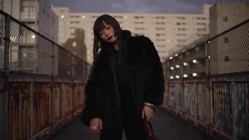 A fashionable Japanese woman in a black fur coat poses with a serious expression on a gritty urban footbridge at dusk