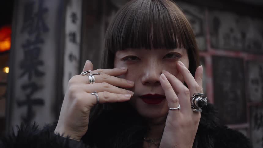 A stylish Asian woman with red lipstick and silver rings holds her face and stares intently into the camera in slow motion on a city street