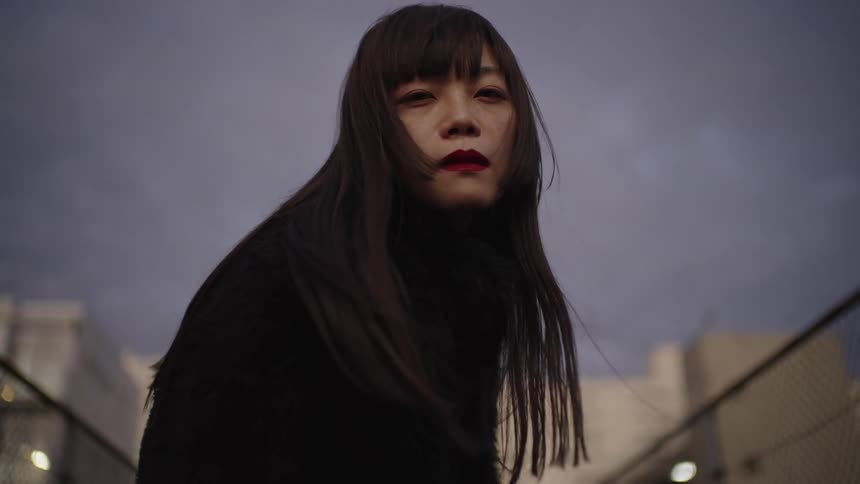 Dramatic low-angle shot of a stylish Asian woman staring intensely into the camera in slow motion on a windy urban evening