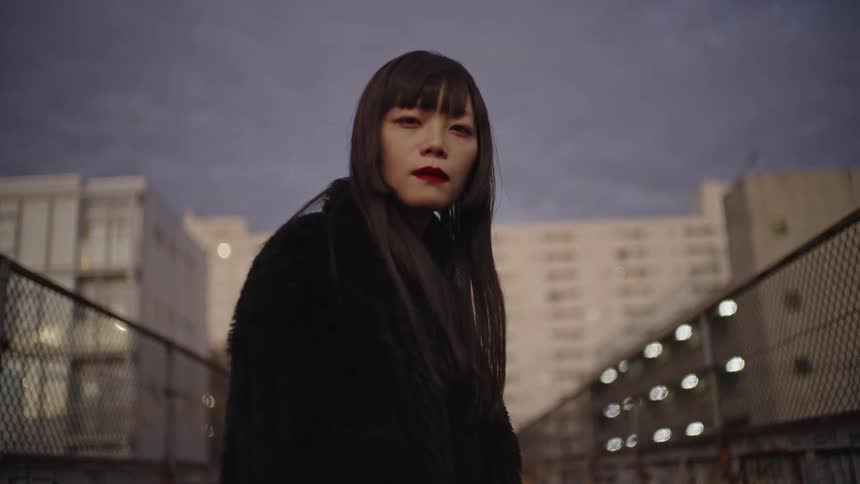 A stylish Japanese woman with red lipstick stares intensely at the camera on an urban walkway at dusk