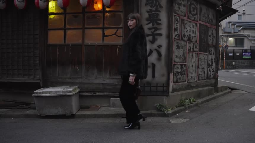 An elegant woman in black walks in slow motion past a traditional Japanese corner bar with glowing lanterns