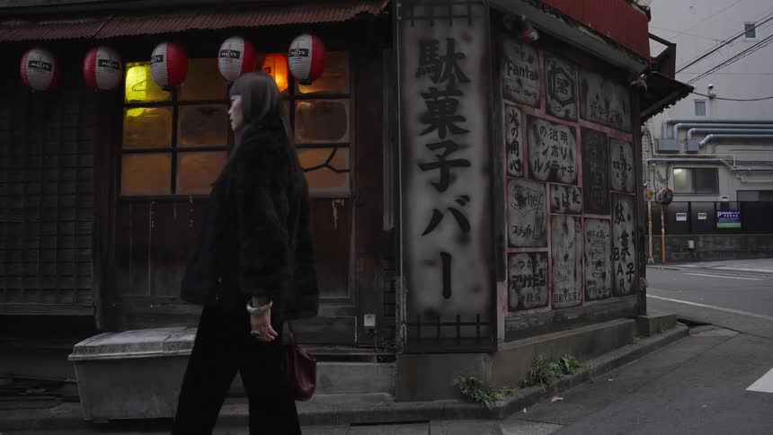 A stylish Japanese woman walks past a traditional corner building with glowing paper lanterns on a city street
