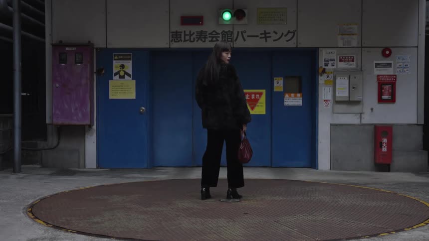 A chic Japanese woman in a black fur coat turns to the camera while standing on a car turntable in a gritty urban parking garage