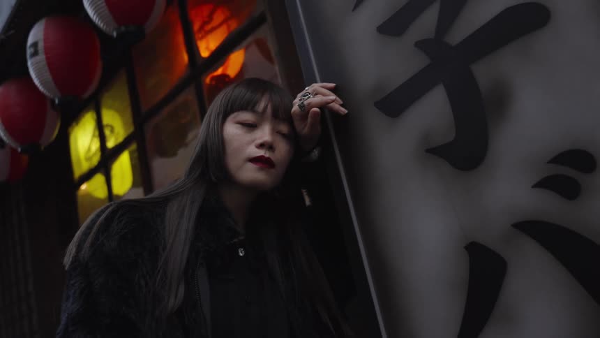 A stylish Japanese woman with long black hair and red lipstick poses with her eyes closed on a city street