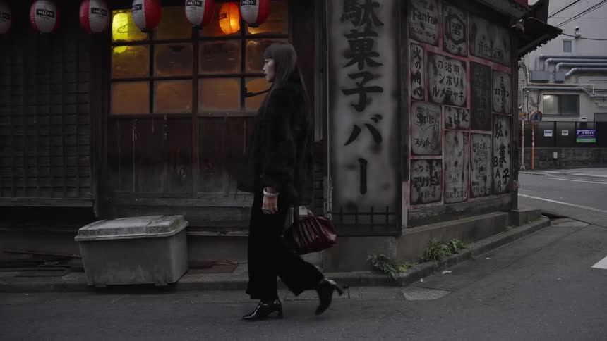 A fashionable woman walks in slow motion past a weathered, traditional Japanese bar with glowing lanterns on a city corner
