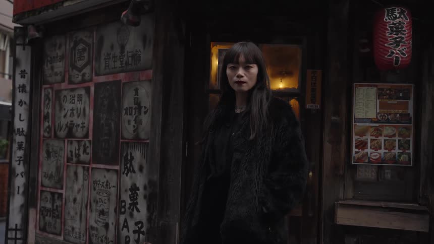 A stylish Japanese woman in a black fur coat poses with a serious expression outside a rustic urban storefront