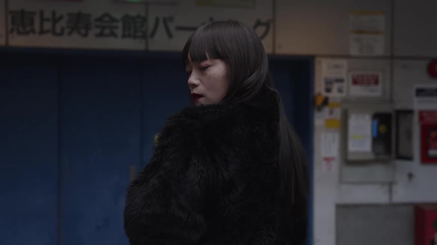 A stylish Japanese woman in a black fur coat looks over her shoulder before walking away in slow motion