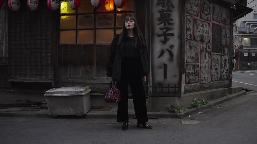 A fashionable young woman in dark clothes stands on a street corner in front of a traditional Japanese bar at dusk