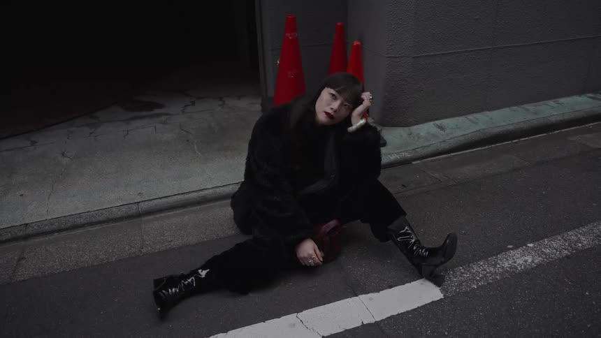 A stylish woman in all black sits on the pavement of a dim urban street, looking directly at the camera