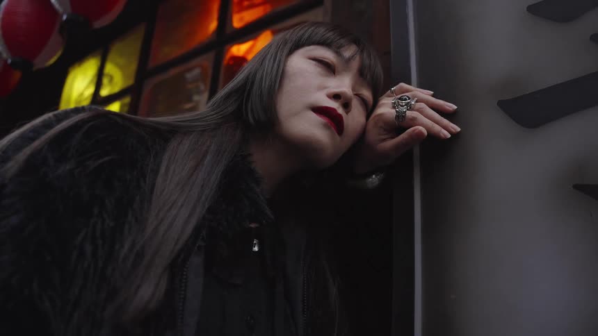 Low-angle shot of a pensive young Asian woman in a black jacket leaning against a wall in a moody, dimly lit urban setting at night